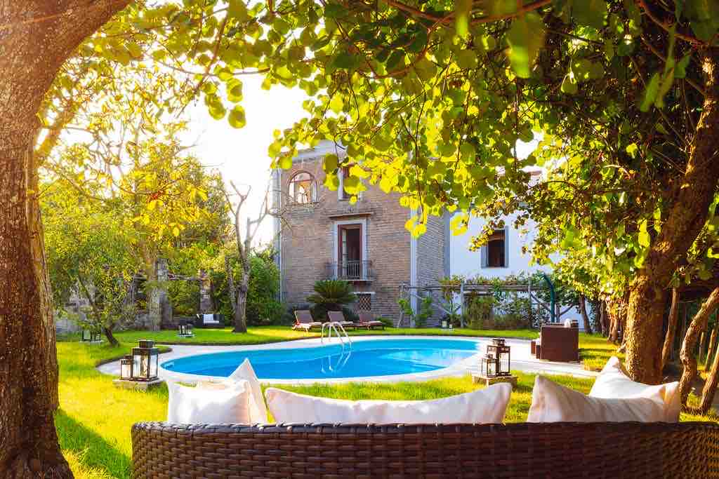 A serene outdoor space features a swimming pool surrounded by lush greenery and trees. Comfortable seating with soft cushions is positioned nearby, inviting relaxation. The elegant building in the background displays classic architectural details, enhancing the tranquil ambiance of the garden.