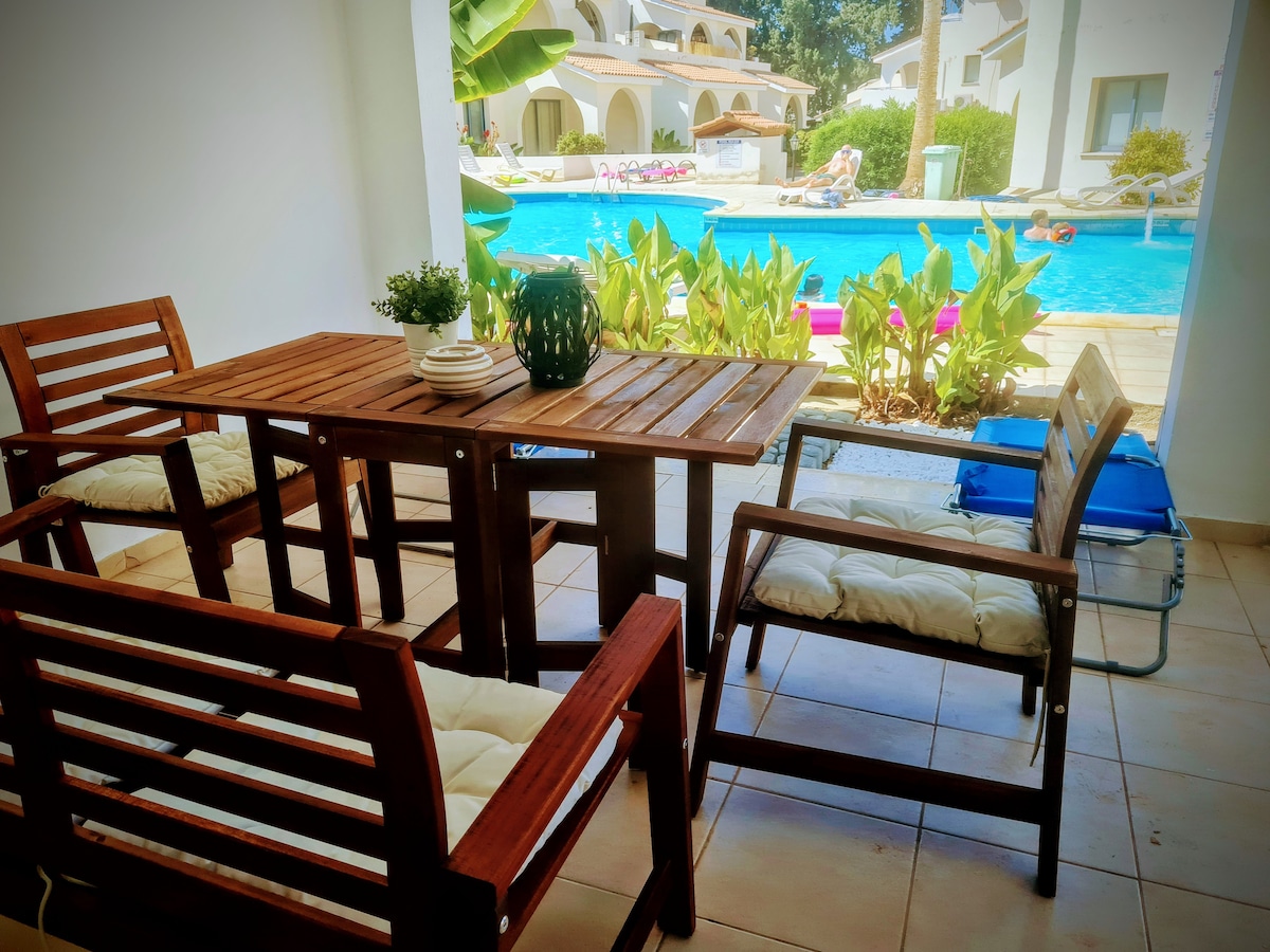 An outdoor seating area is presented, featuring a wooden table surrounded by four chairs. Lush greenery outlines the space, providing a view of the inviting swimming pool in the background, where pool loungers are partially visible.