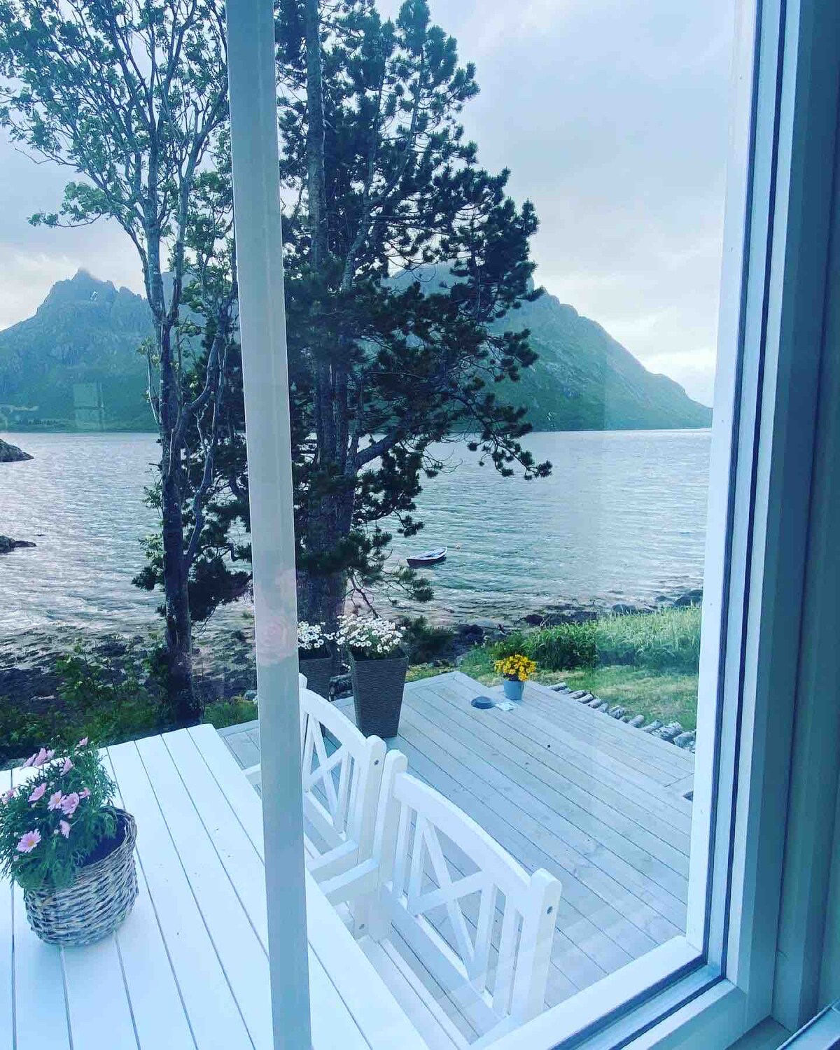 A serene view of the fjord can be seen through large windows, featuring a wooden deck with seating and flower pots. Towering mountains rise in the background, while calm waters reflect the soft light of the overcast sky.