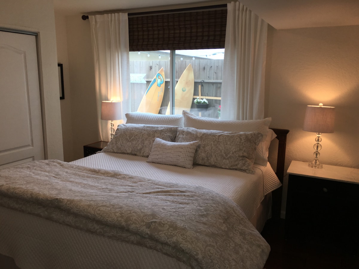 A cozy bedroom features a spacious bed adorned with textured bedding and decorative pillows. Soft lighting from two bedside lamps creates a warm ambiance. Large windows are dressed with sheer curtains, revealing surfboards and a private outdoor area beyond.
