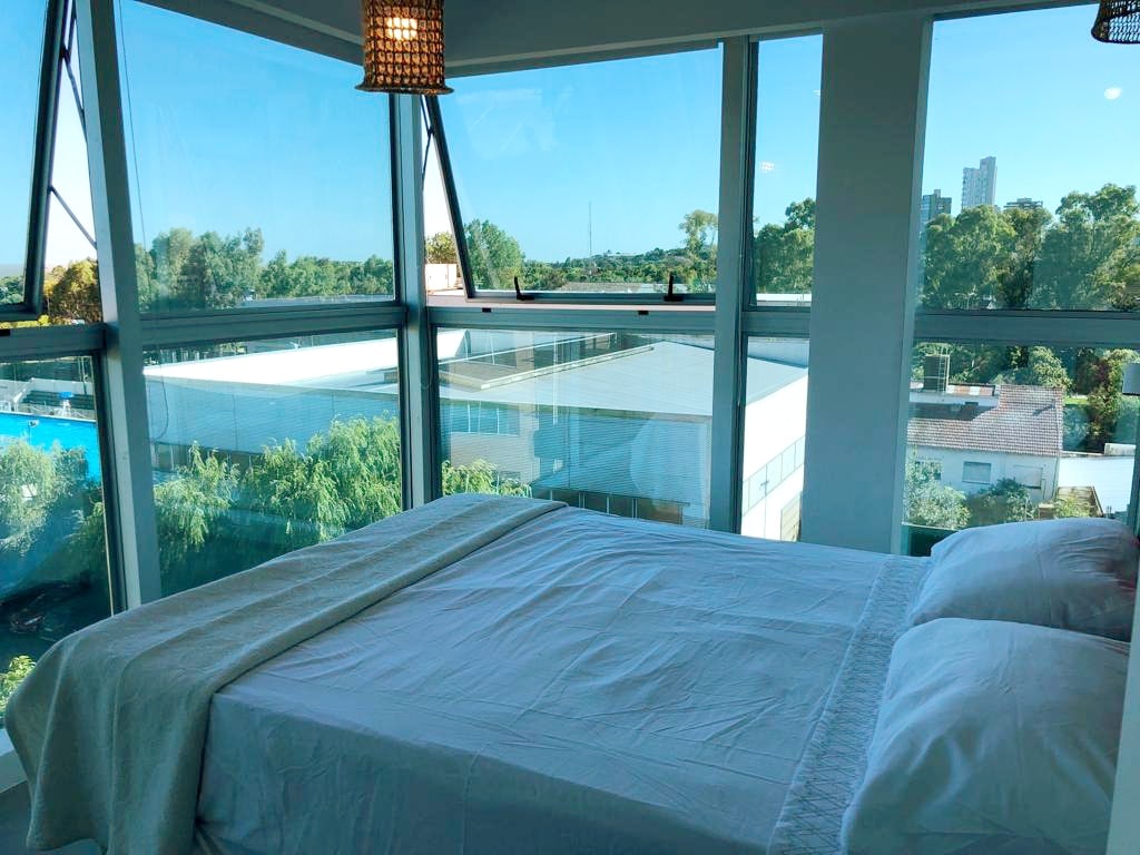 A bright sleeping area features large windows offering a view of the surrounding greenery and buildings. A spacious bed with white linens is centered in the room, while natural light fills the space, highlighting the calm atmosphere.