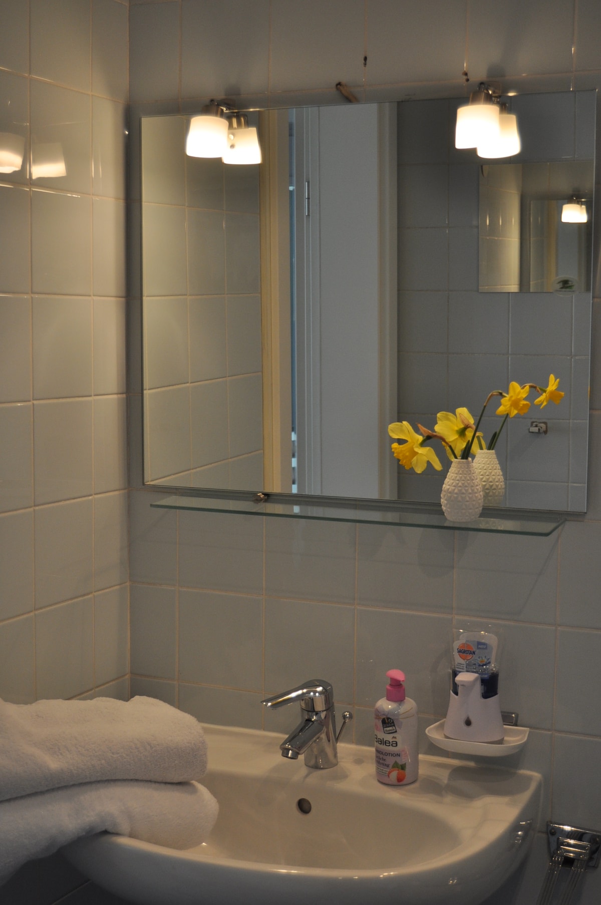 A well-lit bathroom features pale blue tiles and a wall-mounted mirror above the sink. A vase with fresh yellow flowers adds a touch of color, while neatly rolled white towels are positioned by the basin. The shower area is accessible, enhancing functionality.