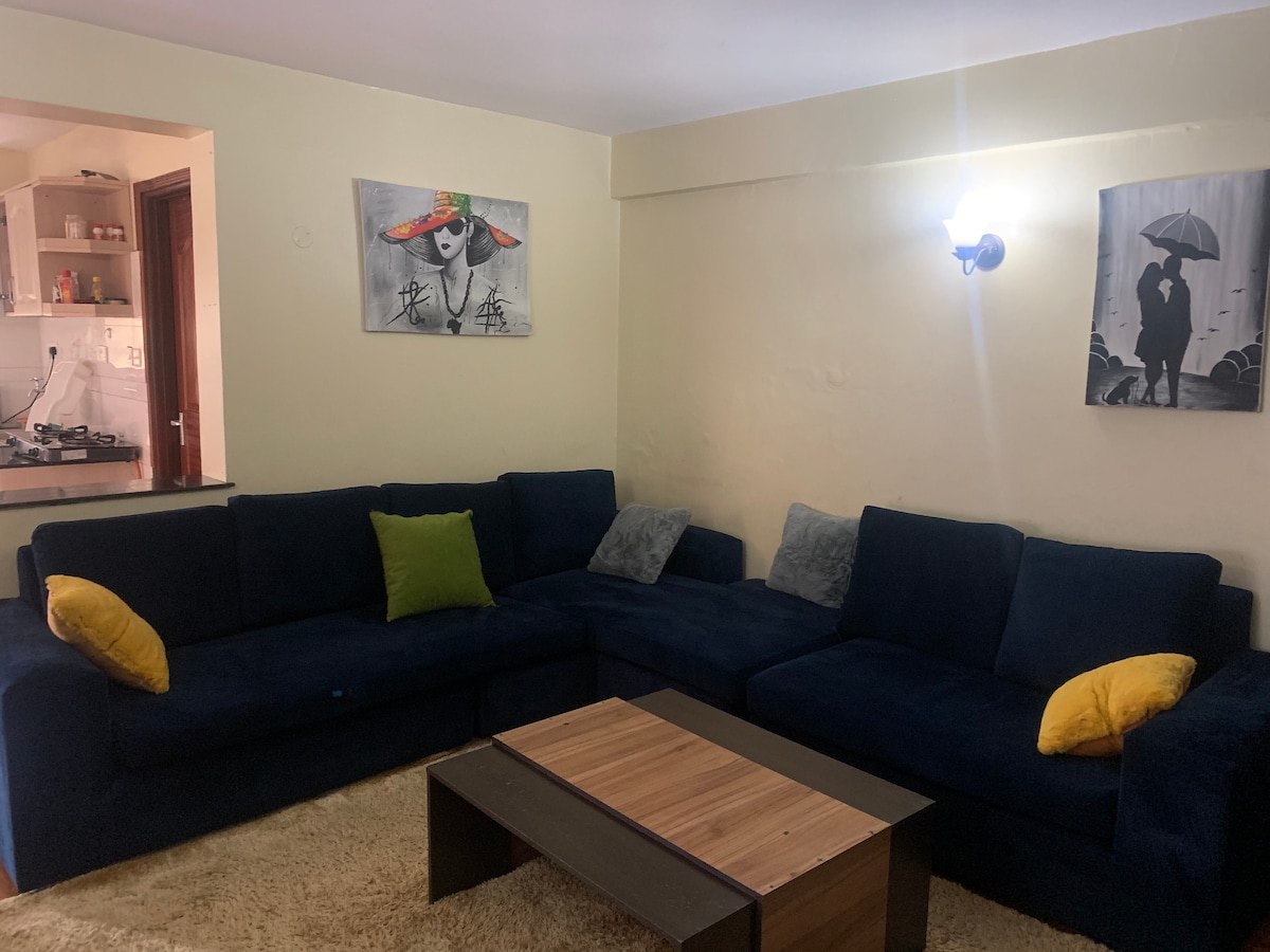 A spacious living area features a large navy-blue sectional sofa adorned with yellow and gray pillows. A wooden coffee table is situated in the center, while light-colored walls are complemented by framed artwork. A glimpse of a kitchenette is visible in the background.