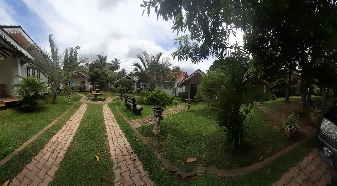 A serene garden area features neatly paved paths lined with greenery and various plants. Several cottages are visible in the background, along with benches and ornamental features scattered throughout. The sky is partly cloudy, enhancing the calm ambiance of the surroundings.