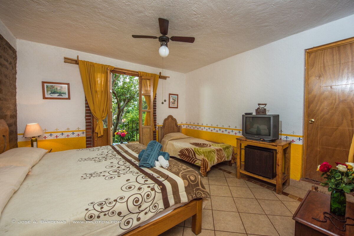The spacious room features two beds with decorative linens, complemented by a wooden furniture set. A window with yellow curtains allows natural light, while a television is positioned beside a wooden cabinet. A ceiling fan is visible, contributing to a comfortable environment.