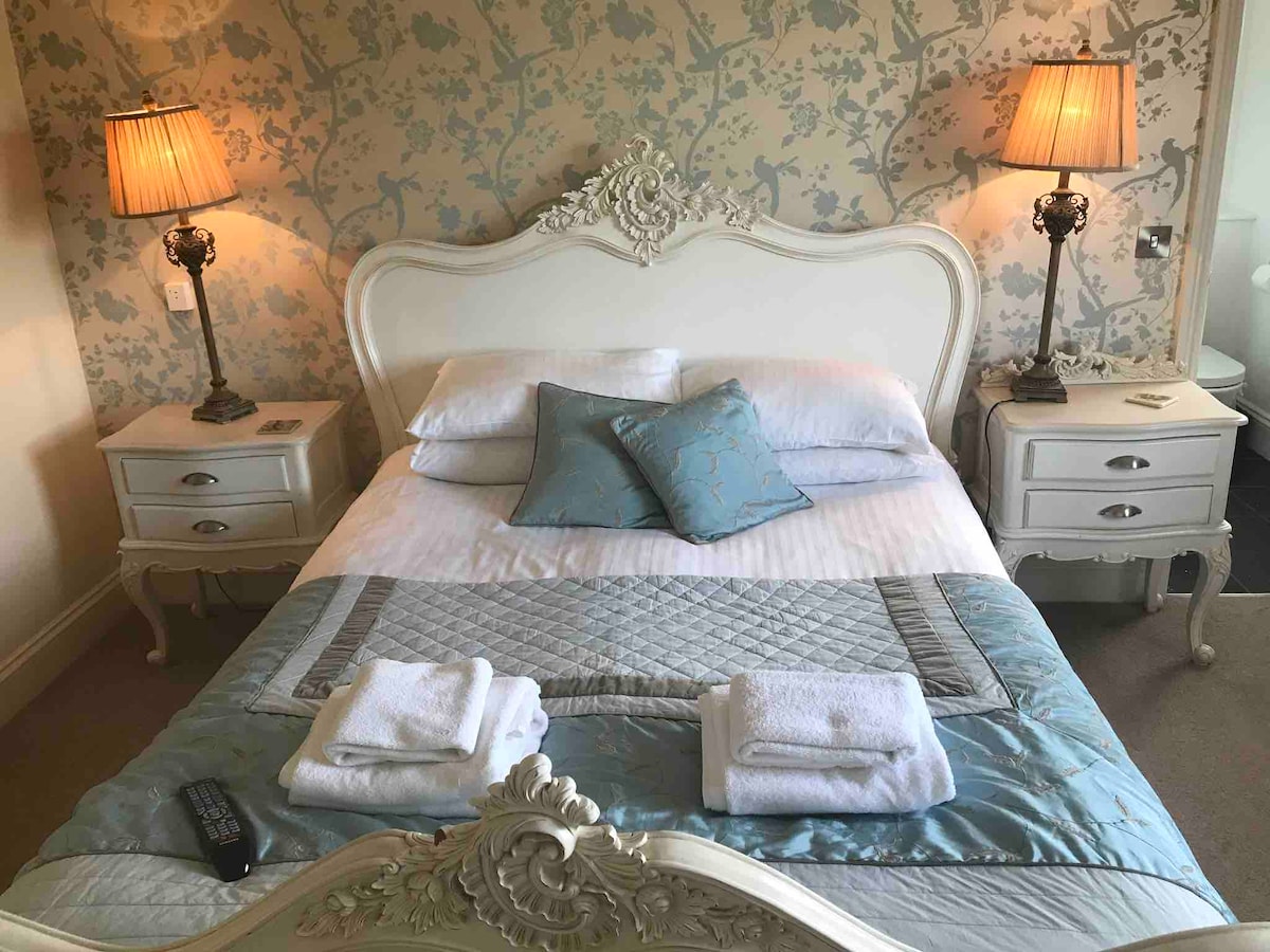 A cozy bedroom setting is presented with a spacious bed adorned in white linens and an elegant blue quilt. Two bedside tables with lamps flank the bed, providing soft lighting. Decorative wallpaper enhances the room's charm, while fresh towels are neatly arranged on the bed.