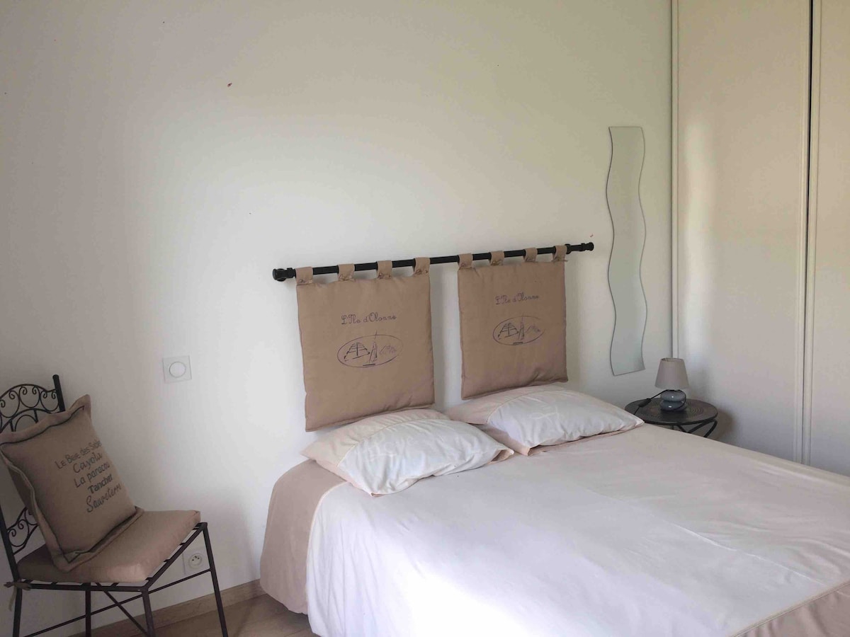 A cozy bedroom is presented with a spacious bed adorned with neutral-toned linens and two decorative pillows. A simple headboard provides support for the pillows. A bedside table holds a lamp, while a chair is positioned nearby, complementing the minimalist design.