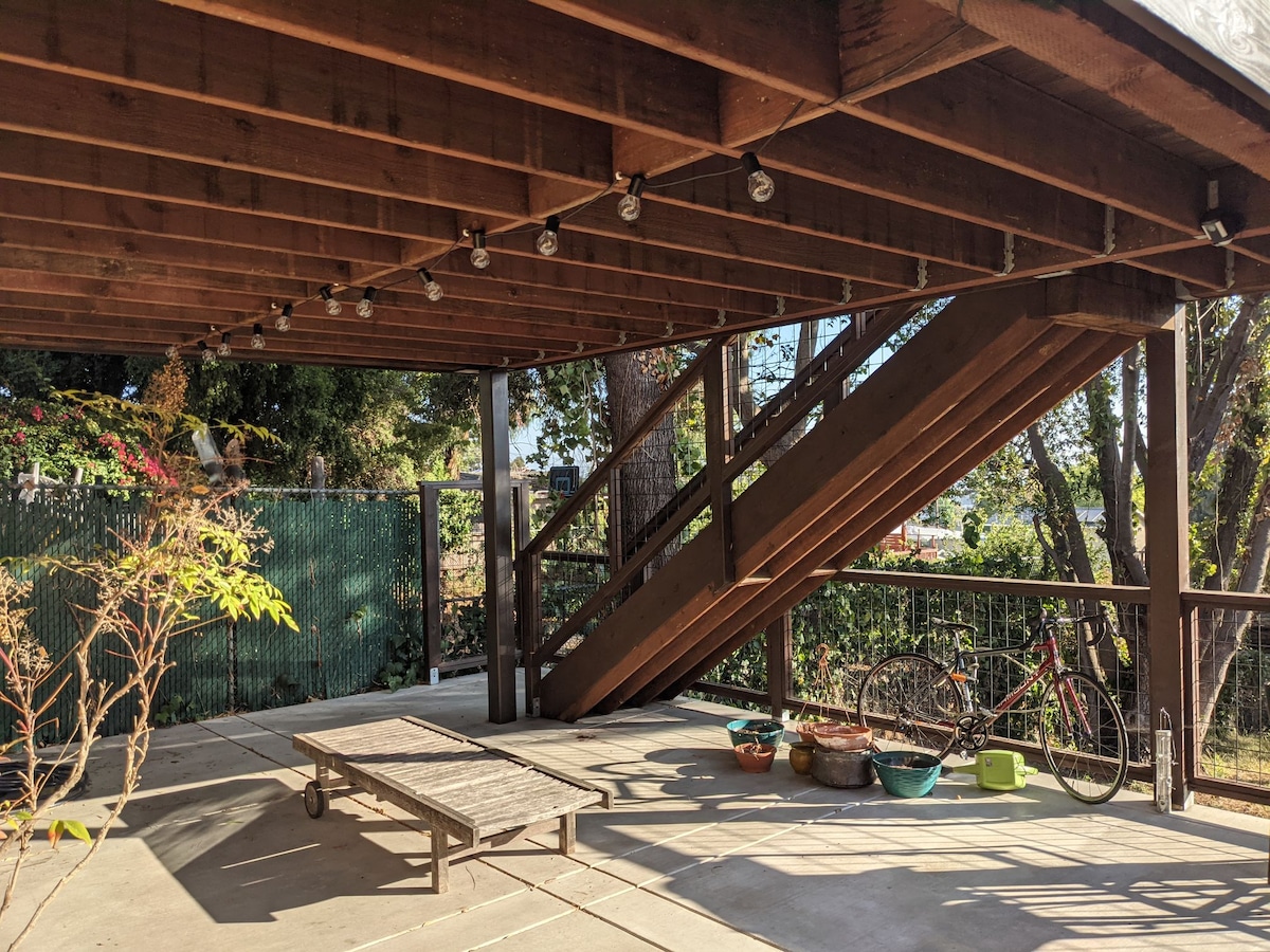An outdoor space is featured, with wooden beams overhead and warm lighting provided by hanging bulbs. A staircase leads to the upper level, and seating arrangements are visible along with potted plants. Bicycles and various containers are positioned in the area.