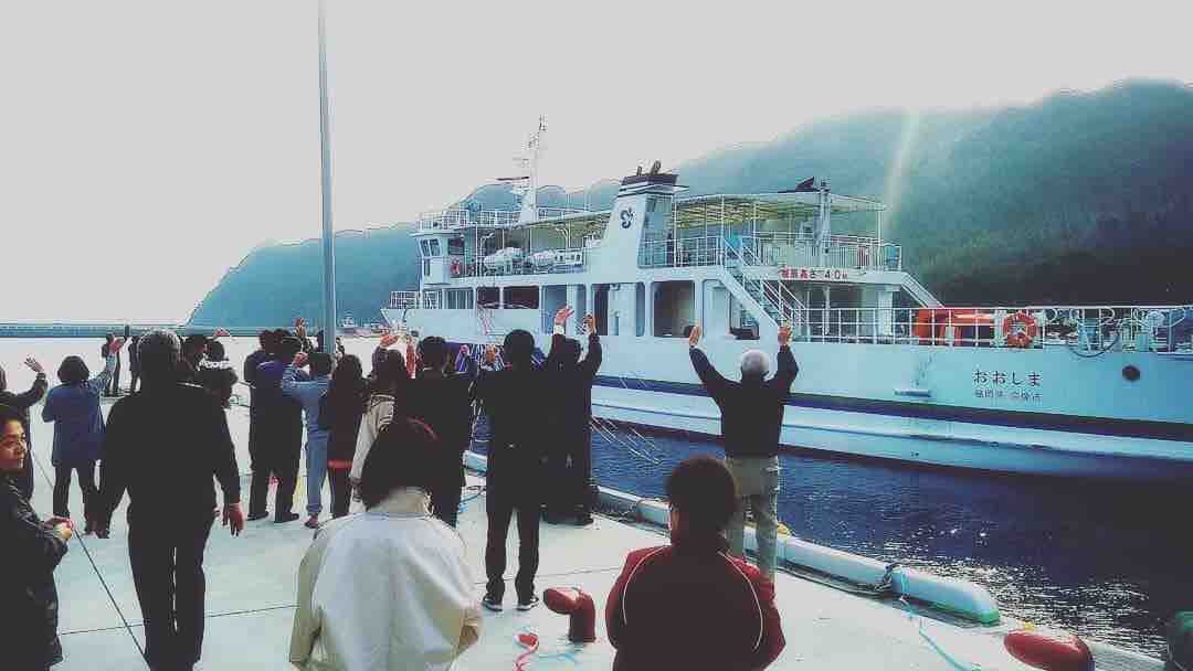 A ferry is seen approaching the dock, with passengers waving from the deck. The scene features a crowd gathered on the dock, against a backdrop of lush, green hills and a clear sky. The atmosphere is lively, capturing a moment of departure.