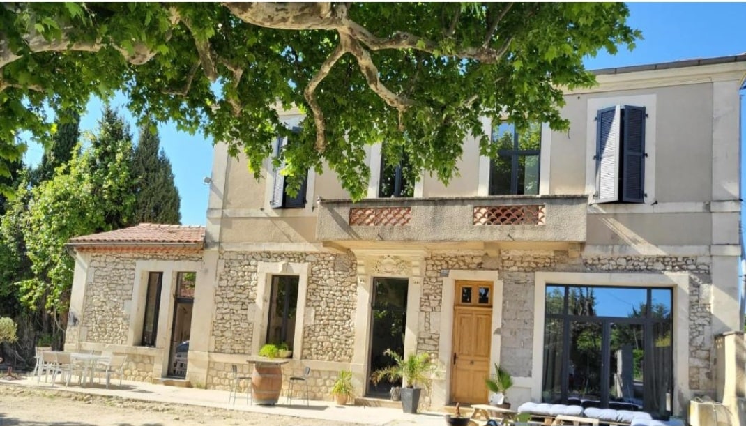 The exterior of the renovated stone house is highlighted by large windows and a welcoming wooden door. A shaded seating area is visible on the right, featuring a white lounge set. Lush greenery surrounds the property, contributing to a serene environment.