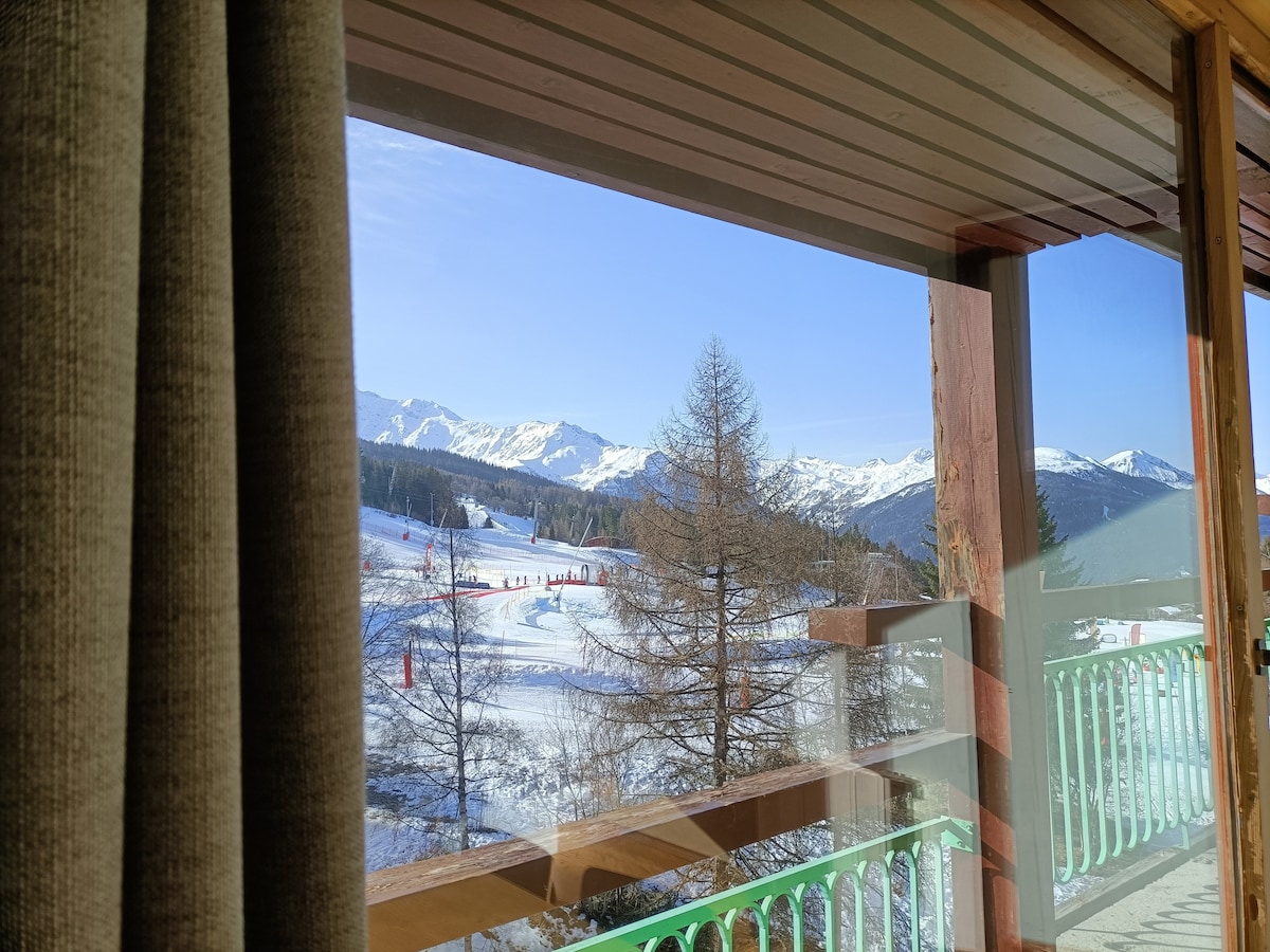 A scenic view is showcased through a large window, displaying snow-covered slopes and majestic mountains. Tall trees are visible in the foreground, and sunlight streams into the space, creating a bright indoor atmosphere.