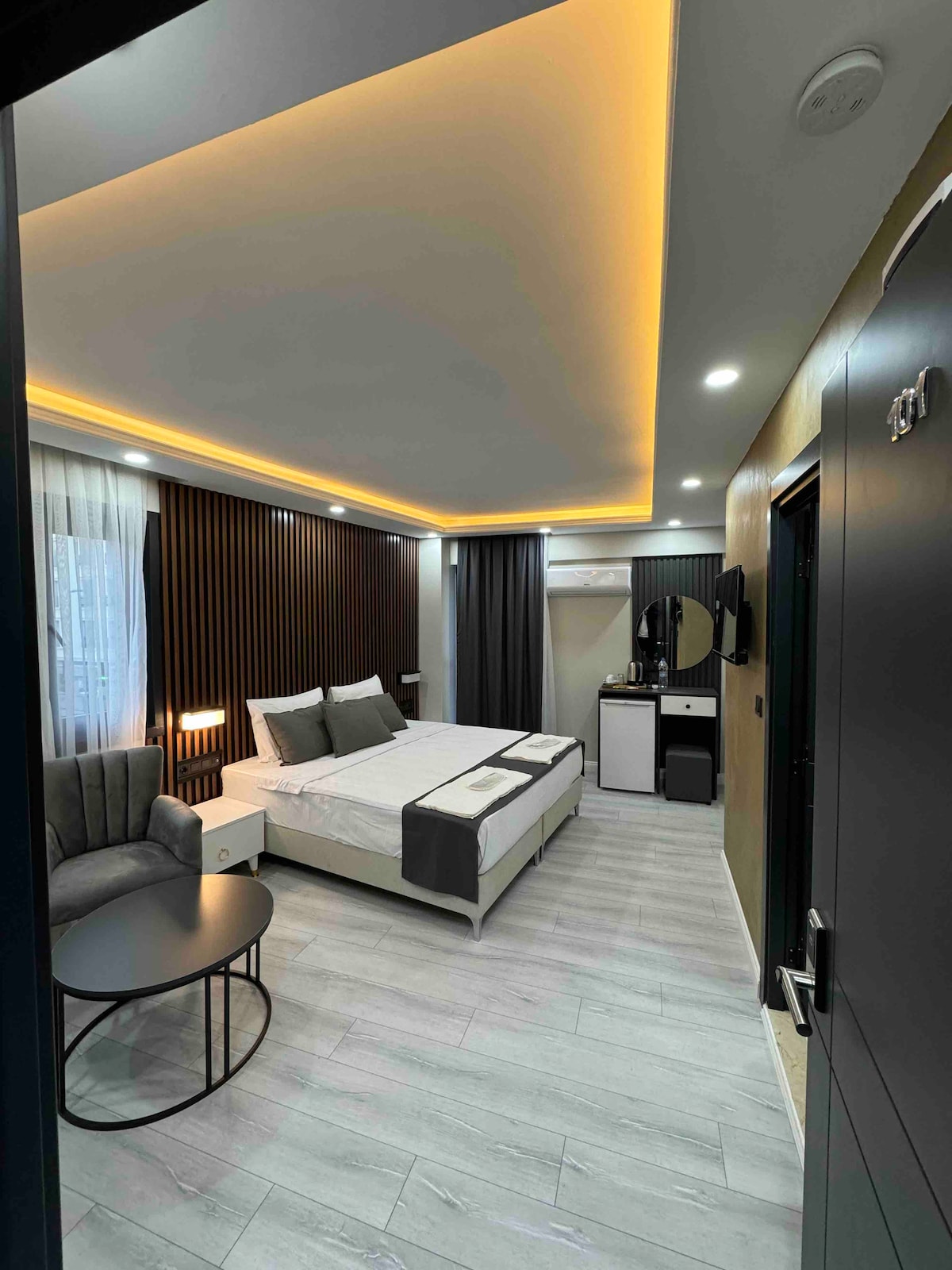 A modern room features a spacious bed centred against a textured wall, complemented by ambient lighting. A comfortable seating area includes a grey armchair and a round table. A mini-fridge and mirror are visible, contributing to the room's functionality.