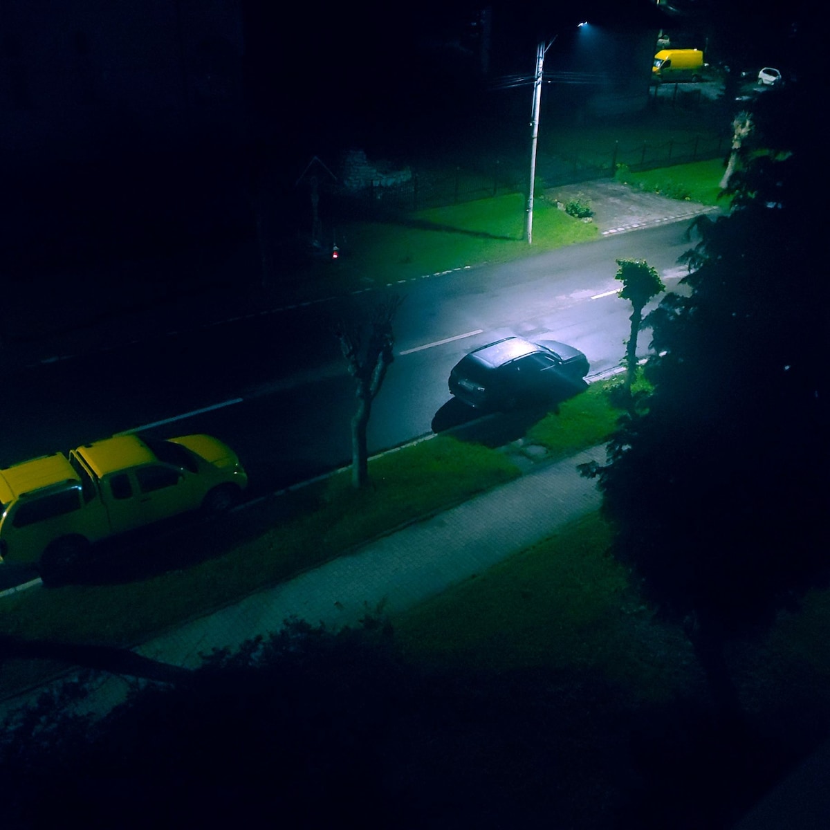 A nighttime street scene is illuminated by streetlights, revealing a wet road glistening in the dark. A single car is parked along the curb, while yellow vehicles are visible in the background, suggesting a quiet neighborhood atmosphere.