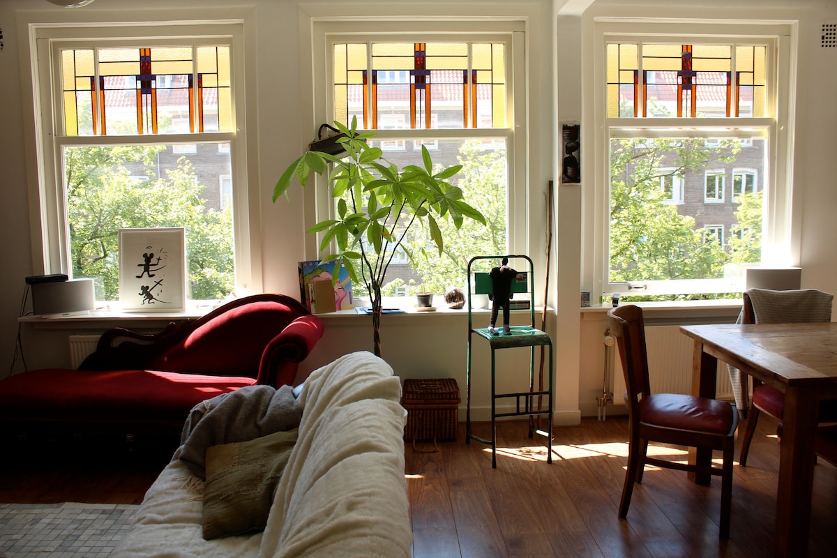 A cozy lounge area features large stained glass windows, allowing natural light to brighten the space. A plush red sofa faces towards a wooden dining table and chairs. Potted plants add greenery to the room, enhancing the inviting atmosphere.