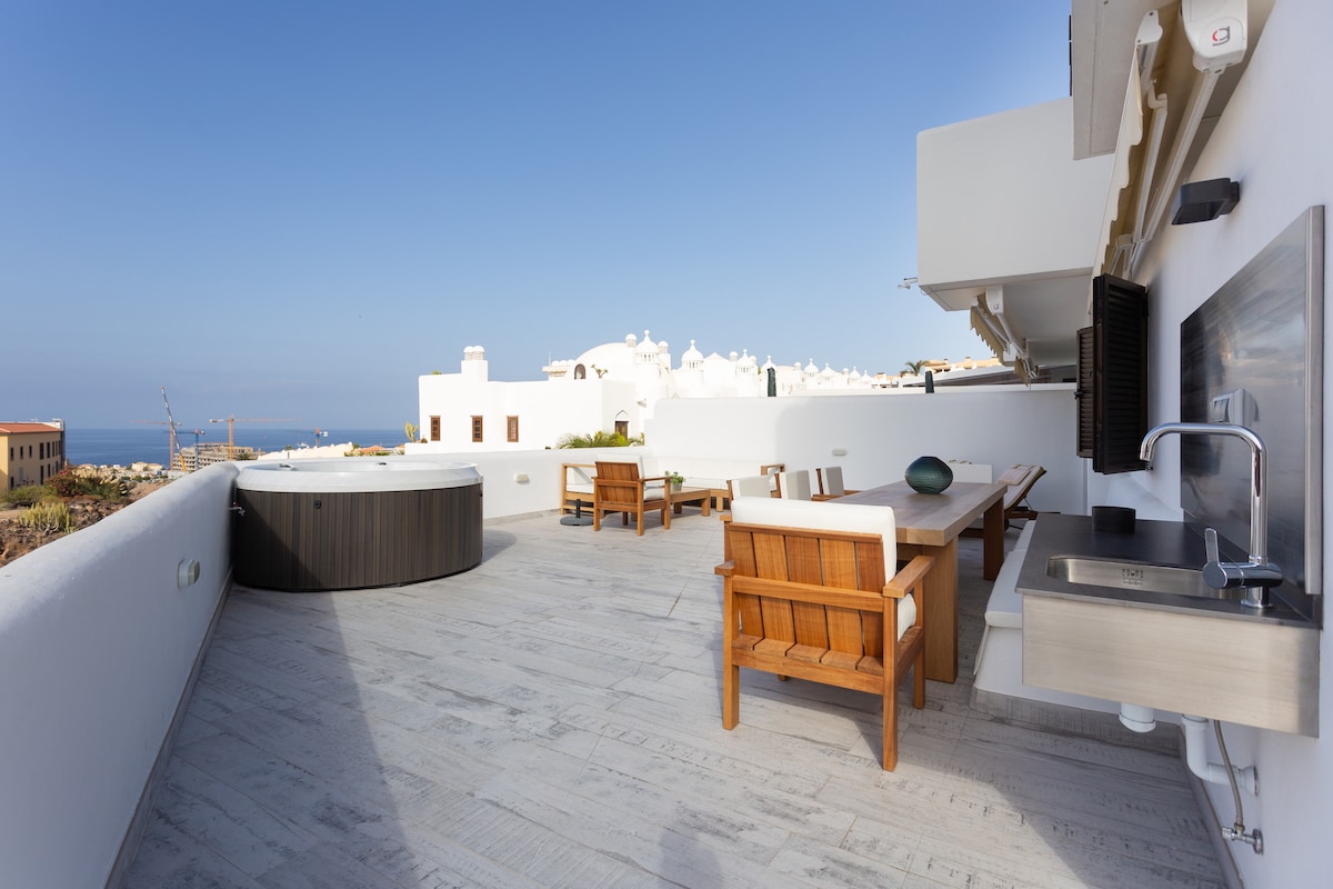 A spacious terrace features a private jacuzzi and a dining table with wooden chairs. An outdoor kitchen area is visible, complemented by a scenic ocean view and a clear blue sky. The flooring is made of light-colored tiles, enhancing the open atmosphere.