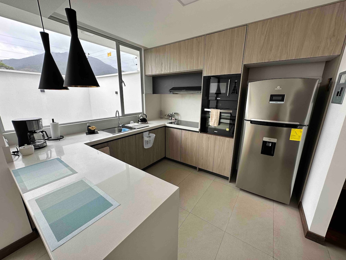 The kitchen features modern cabinetry with a clean design, showcasing stainless steel appliances, including a refrigerator. A sleek countertop is complemented by a window providing natural light and views of the outdoors. Two pendant lights hang over a spacious area for meal preparation.
