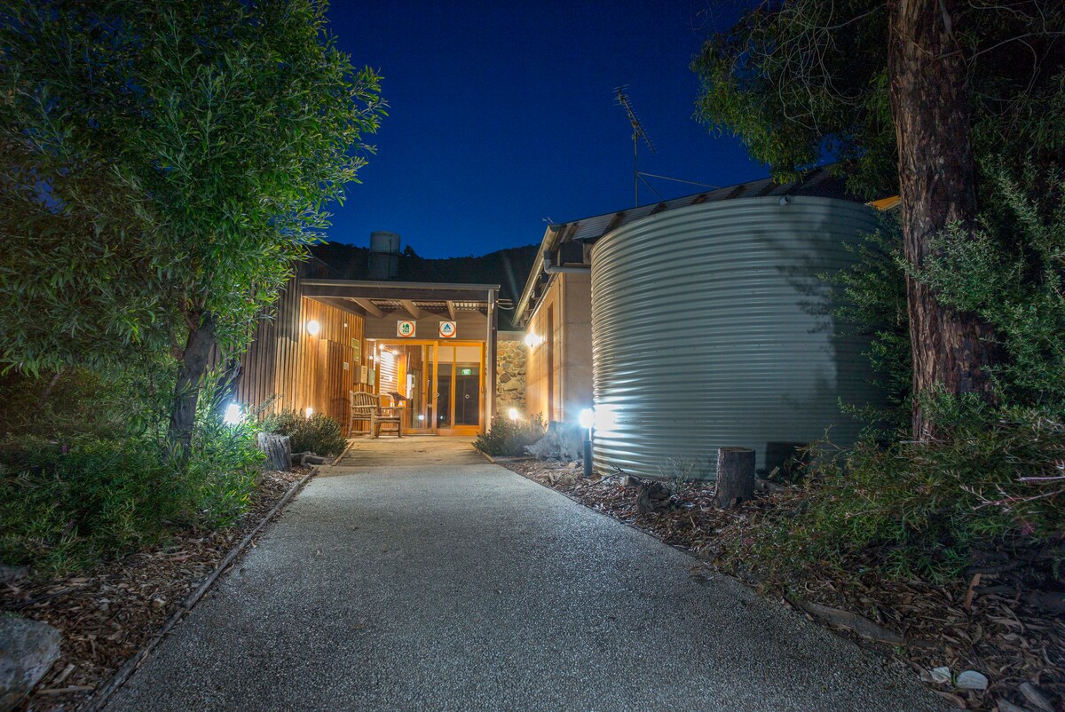 A well-lit pathway leads to the entrance of the hostel, surrounded by lush greenery. The entrance showcases large glass doors that invite guests inside, while a round rainwater tank stands alongside the building, contributing to the natural setting.