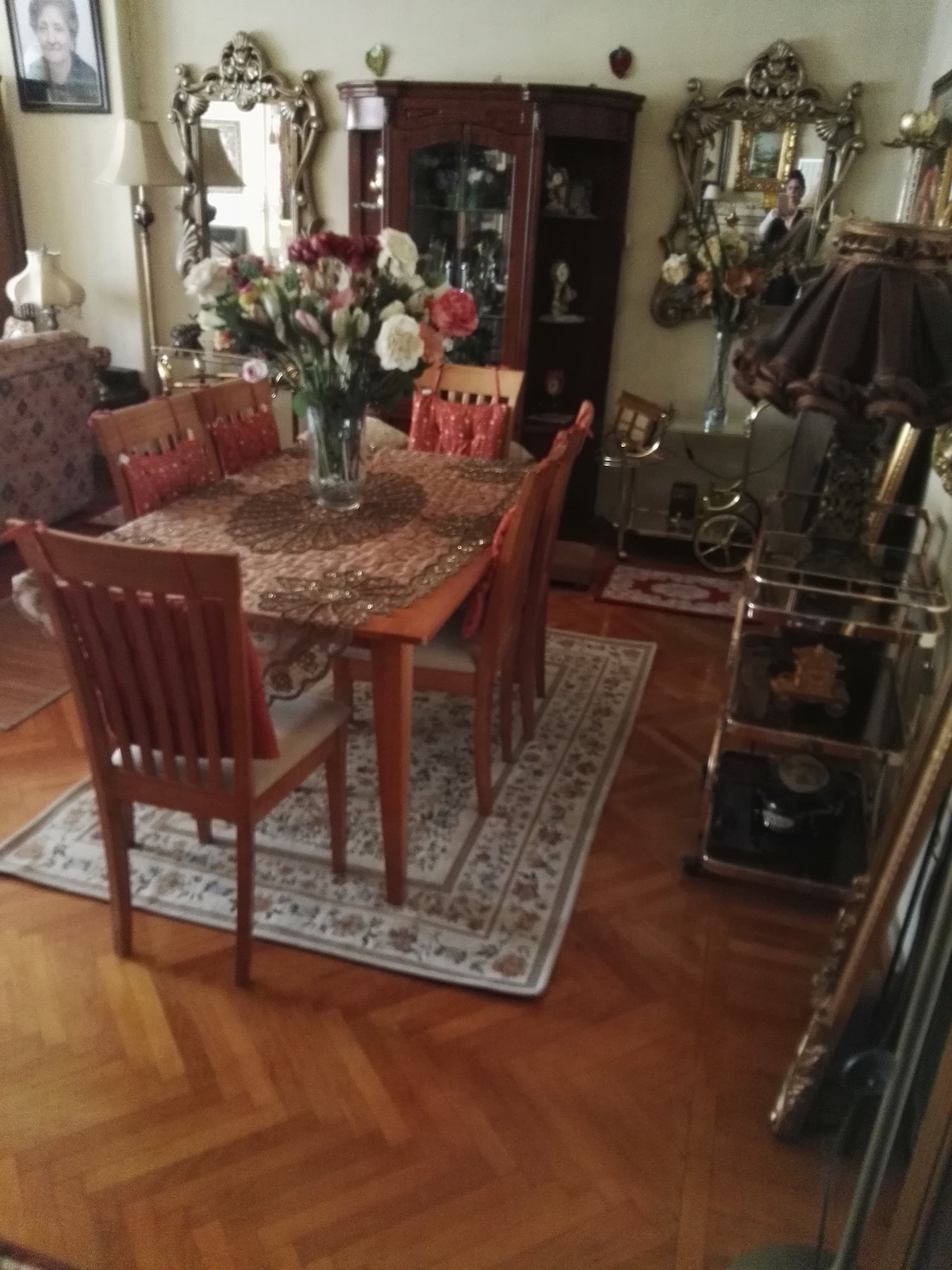A dining area features a wooden table surrounded by six chairs, complemented by a floral centerpiece. A patterned area rug covers the hardwood floor, with various decorative items visible on nearby shelves and tables, reflecting a warm and welcoming ambiance.