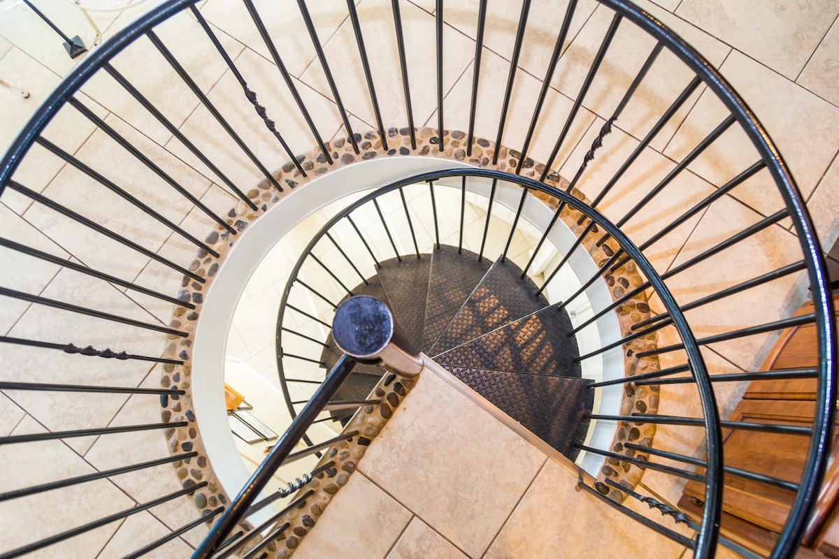 A circular staircase with a black wrought iron railing spirals downwards. The steps are made of light-colored tile, and a textured black runner runs along the center. Natural light partially illuminates the staircase, highlighting its elegant curves.