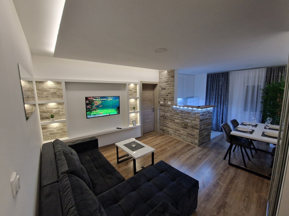 A modern living space features a comfortable dark sofa facing a mounted television. Soft lighting enhances the room, and a dining area with a large table is visible in the background, surrounded by elegant chairs. Natural textures are added with a stone accent wall.