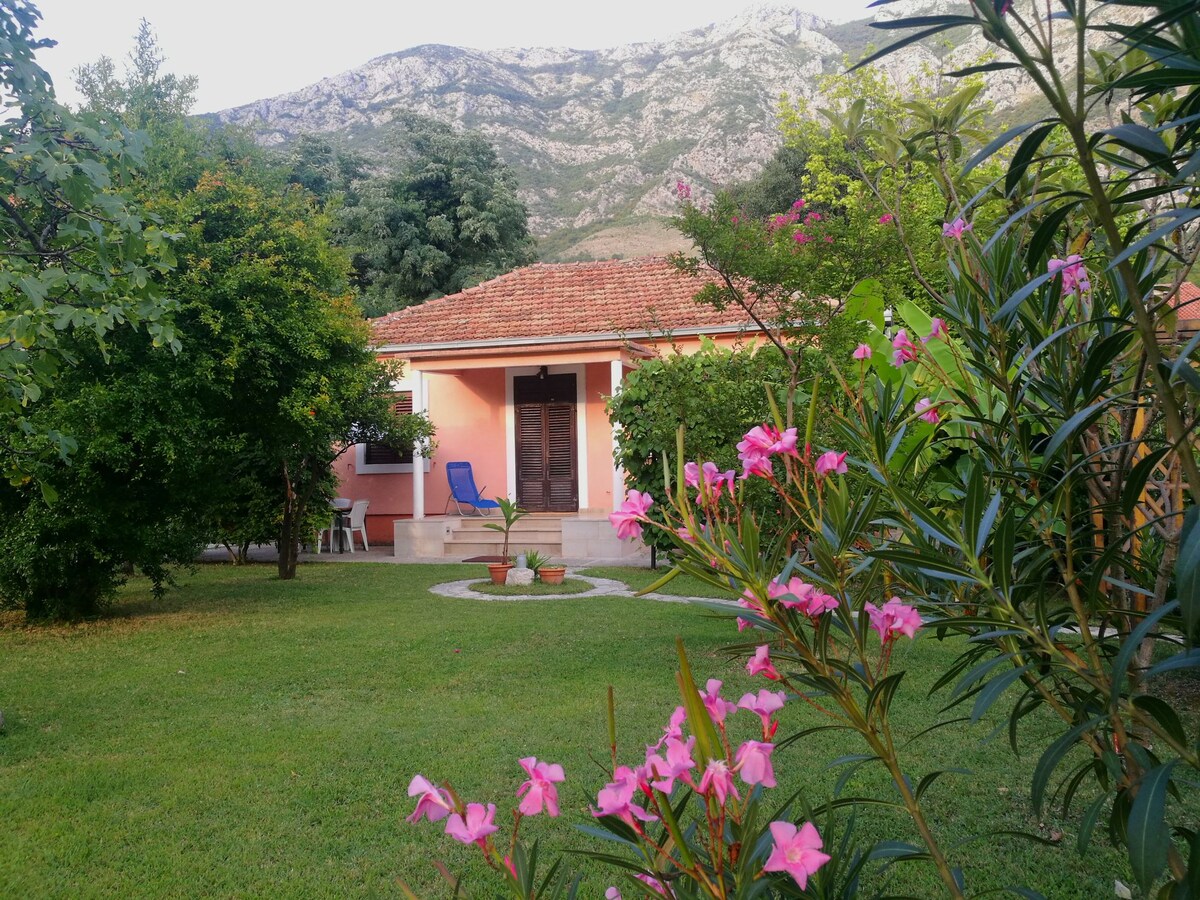 A charming house with a tiled roof is surrounded by a well-maintained yard featuring lush green grass and blooming flowers. A paved path leads to the entrance, where a seating area is visible. Majestic mountains stand in the background, enhancing the natural setting.