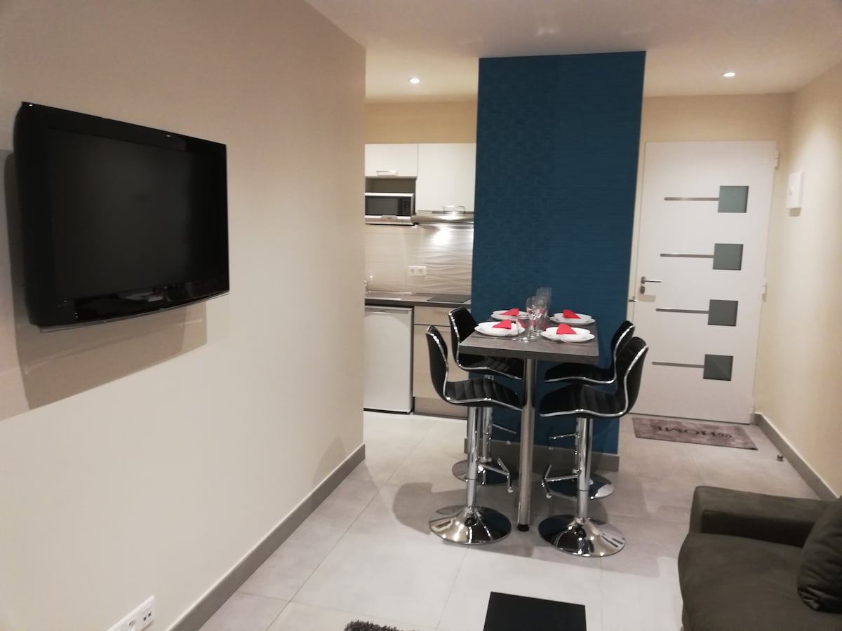 A modern dining area is shown with a round table set for four, surrounded by sleek black chairs. The wall features a textured blue accent, while a flat-screen TV is mounted nearby. The open kitchen is visible in the background, equipped with contemporary appliances.