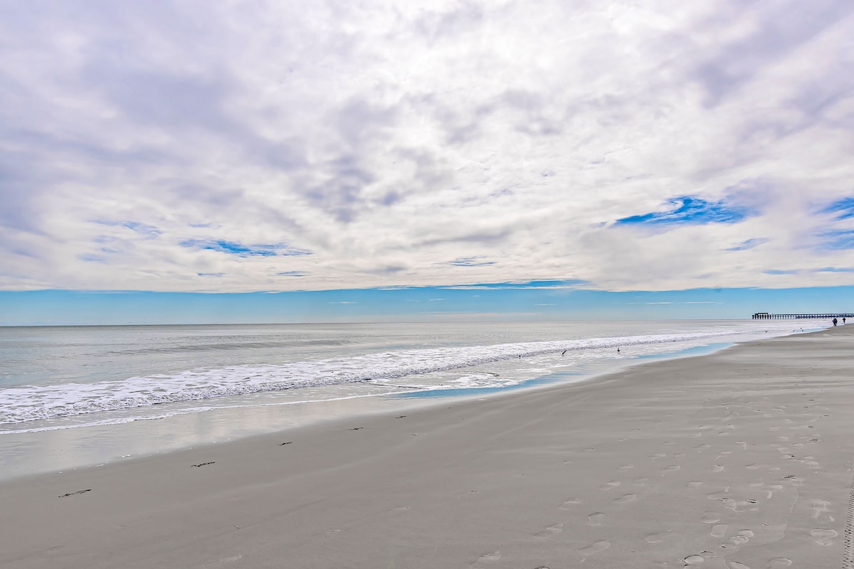 An expansive view of a serene beach scene is displayed, featuring gentle waves lapping at a smooth sandy shoreline. The sky is partly cloudy with soft hues of blue and white. A pier can be seen in the distance, adding depth to the landscape.