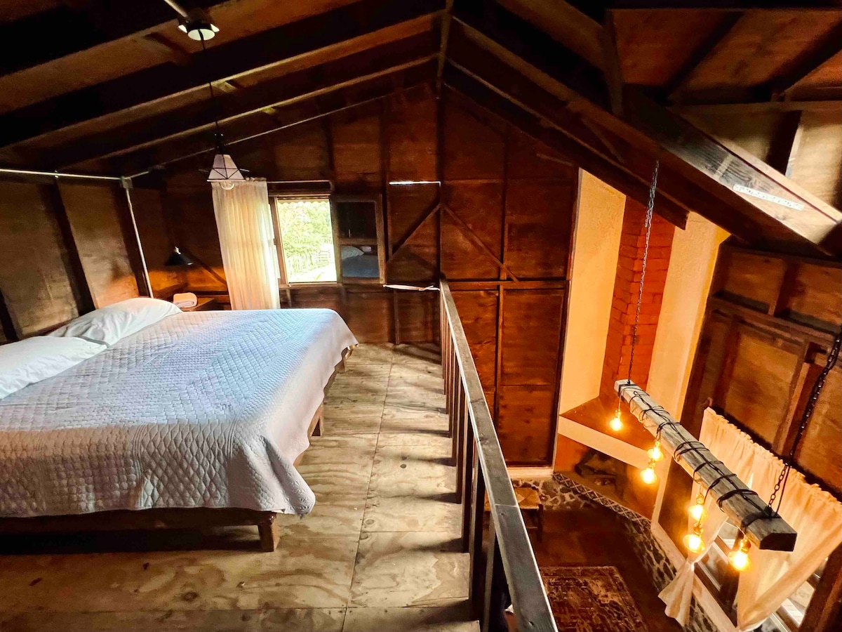 A wooden loft features a spacious bed with a light quilt, complemented by warm wood paneling. Soft light filters through sheer curtains, while a unique hanging light fixture adds ambiance. A staircase leads down to the lower level, enhancing the open design.