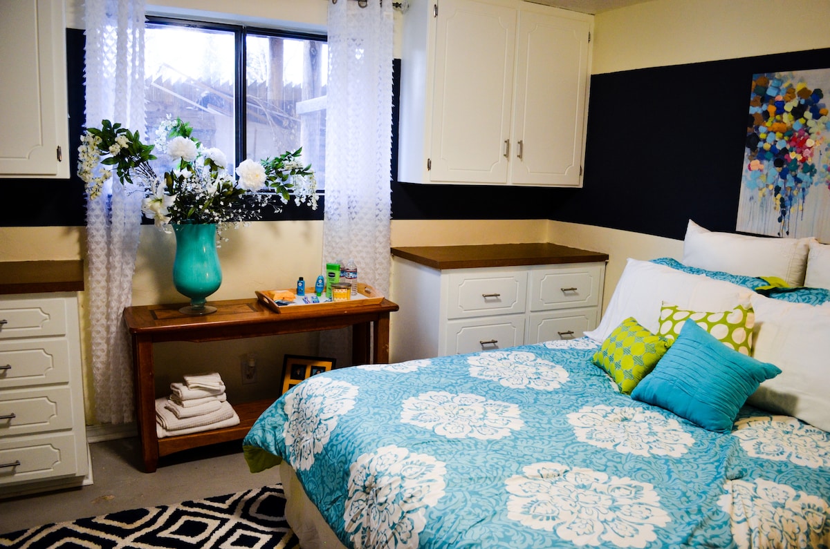 A bright sleeping area is furnished with a spacious bed covered in a blue and white patterned quilt. A worktable sits nearby, adorned with toiletries and a decorative vase filled with flowers. Clear curtains allow natural light to illuminate the room.
