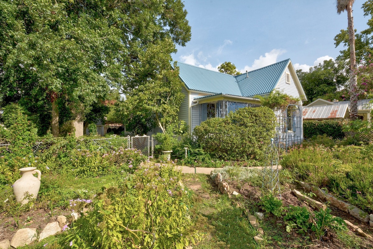 A historic cottage is set amongst lush gardens, featuring a well-maintained path leading to the entrance. Surrounding greenery includes diverse plants and flowering shrubs, complemented by a charming blue metal roof. A decorative clay pot is positioned in the foreground, enhancing the natural setting.