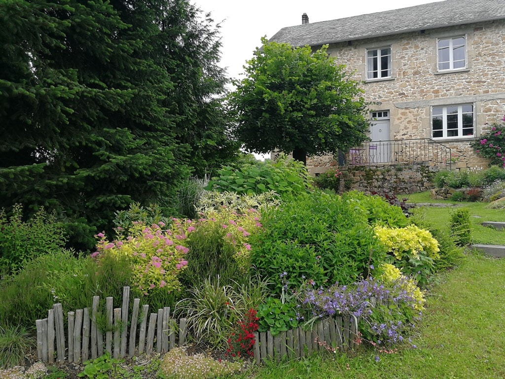 A charming stone house is surrounded by a lush garden featuring a variety of colorful plants, flowers, and greenery. The garden is bordered by wooden fencing, and tall trees provide a natural backdrop, enhancing the tranquil outdoor setting.