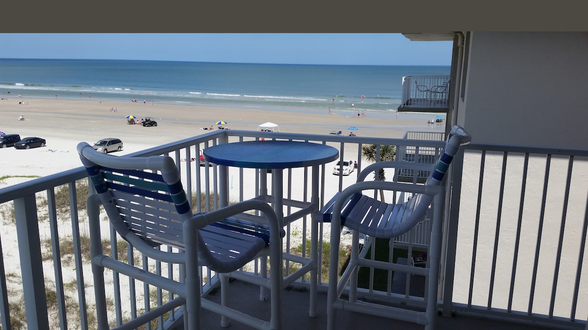 A balcony with a round table and two chairs offers unobstructed views of the beach and ocean. Sandy shores and colorful umbrellas are visible below, while vehicles are parked along the coastline. The scene captures a serene coastal environment.