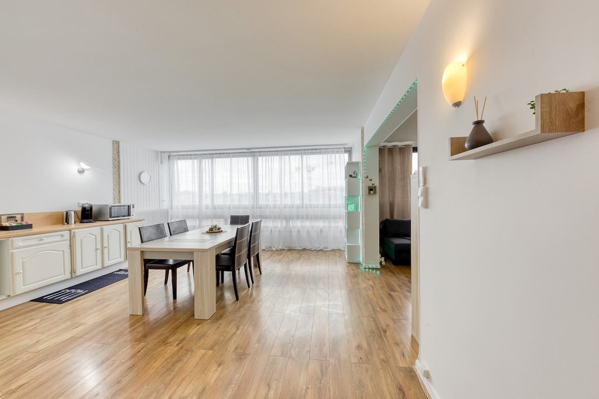 A well-lit open living area is depicted, featuring a dining table surrounded by four dark chairs. Large windows allow natural light to fill the space, while light-colored cabinets enhance the modern kitchen area. A cozy seating area is visible in the background.