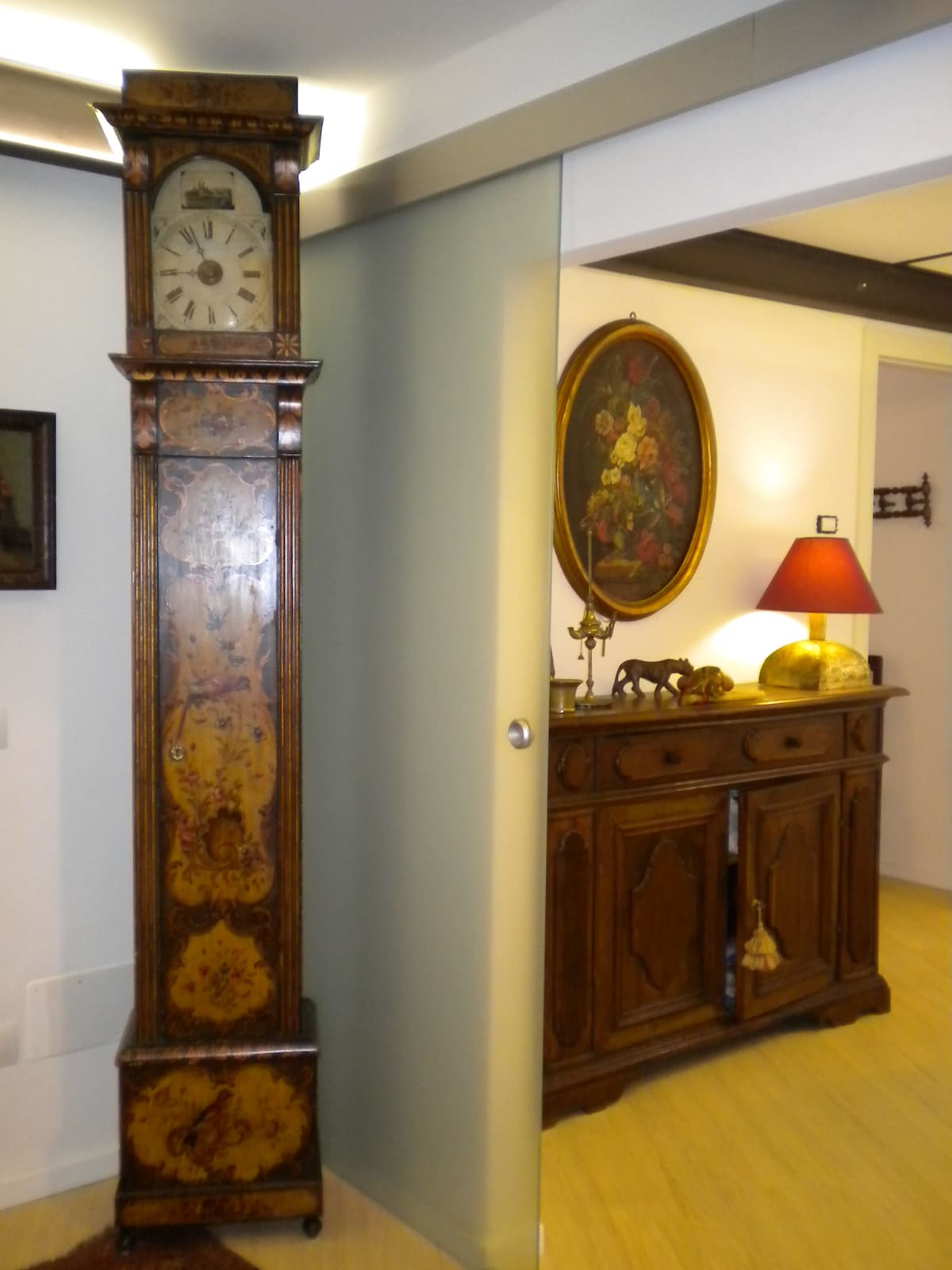 A tall, ornate clock stands against a wall, featuring intricate floral designs and a visible clock face. To the right, a wooden cabinet exhibits classic craftsmanship, complemented by a table lamp with a warm glow and an ornate painting above it, enhancing the elegant atmosphere.