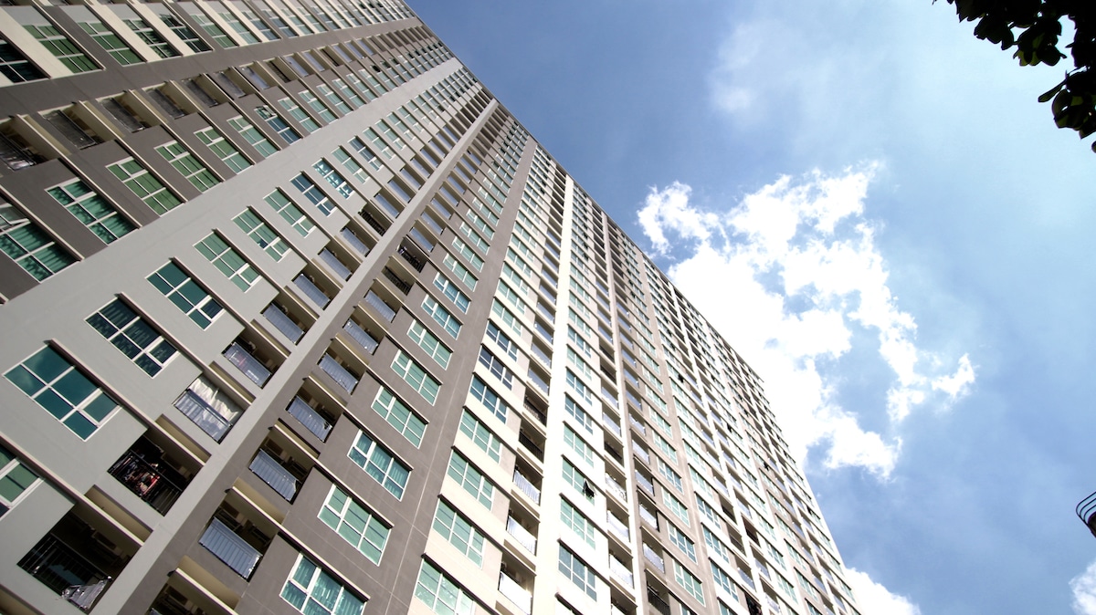 The image captures a tall residential building reaching towards a bright blue sky, with white clouds scattered. The façade is marked by numerous windows, showcasing a modern architectural style, and the structure is displayed from a low angle, emphasizing its height.