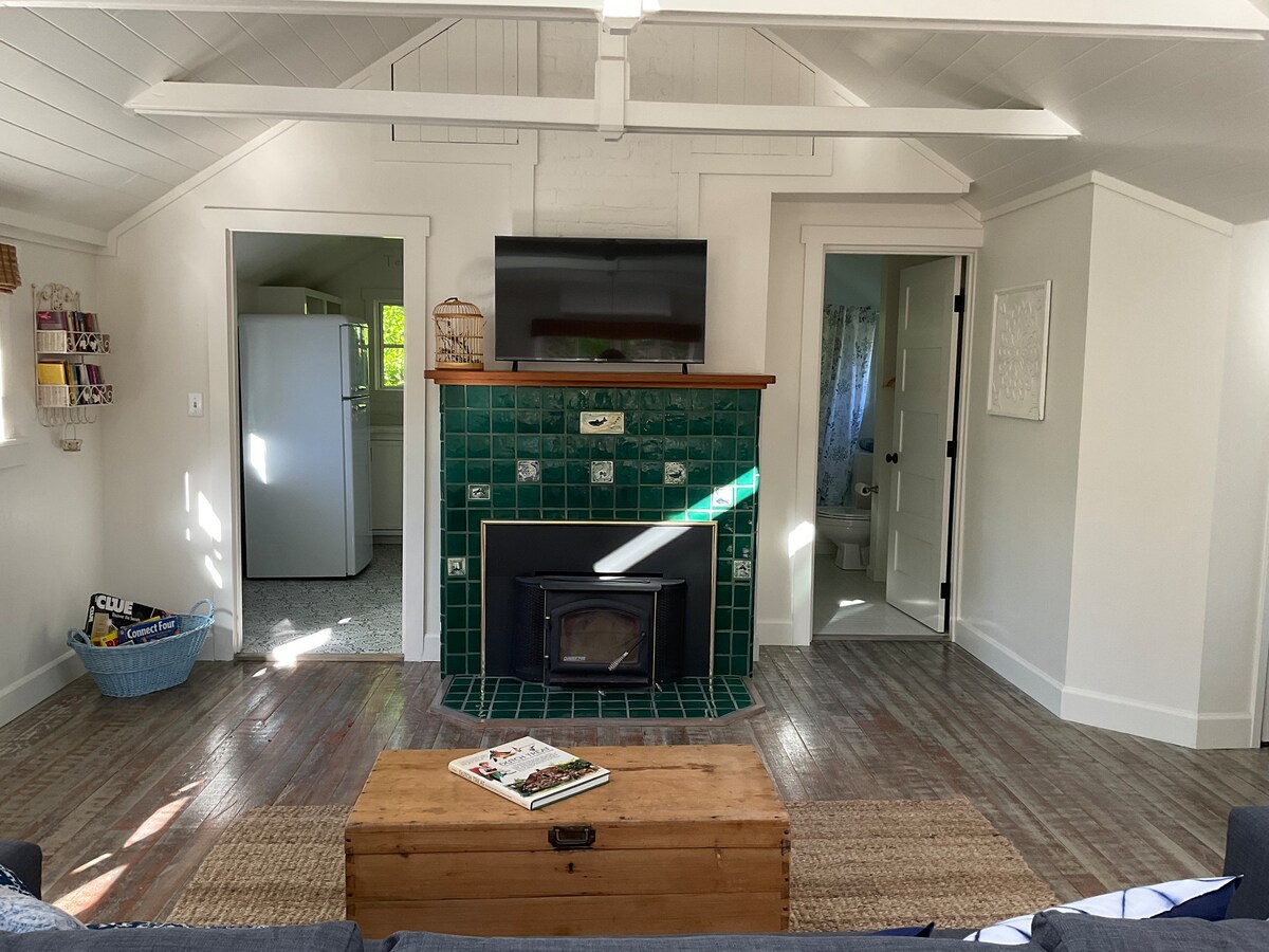 A cozy living room area features a wood-burning fireplace with a green tiled surround and a mounted television above. Natural light enters through the windows, illuminating the wooden floors. A coffee table rests in front of a navy sofa, flanked by two doorways leading to other spaces.