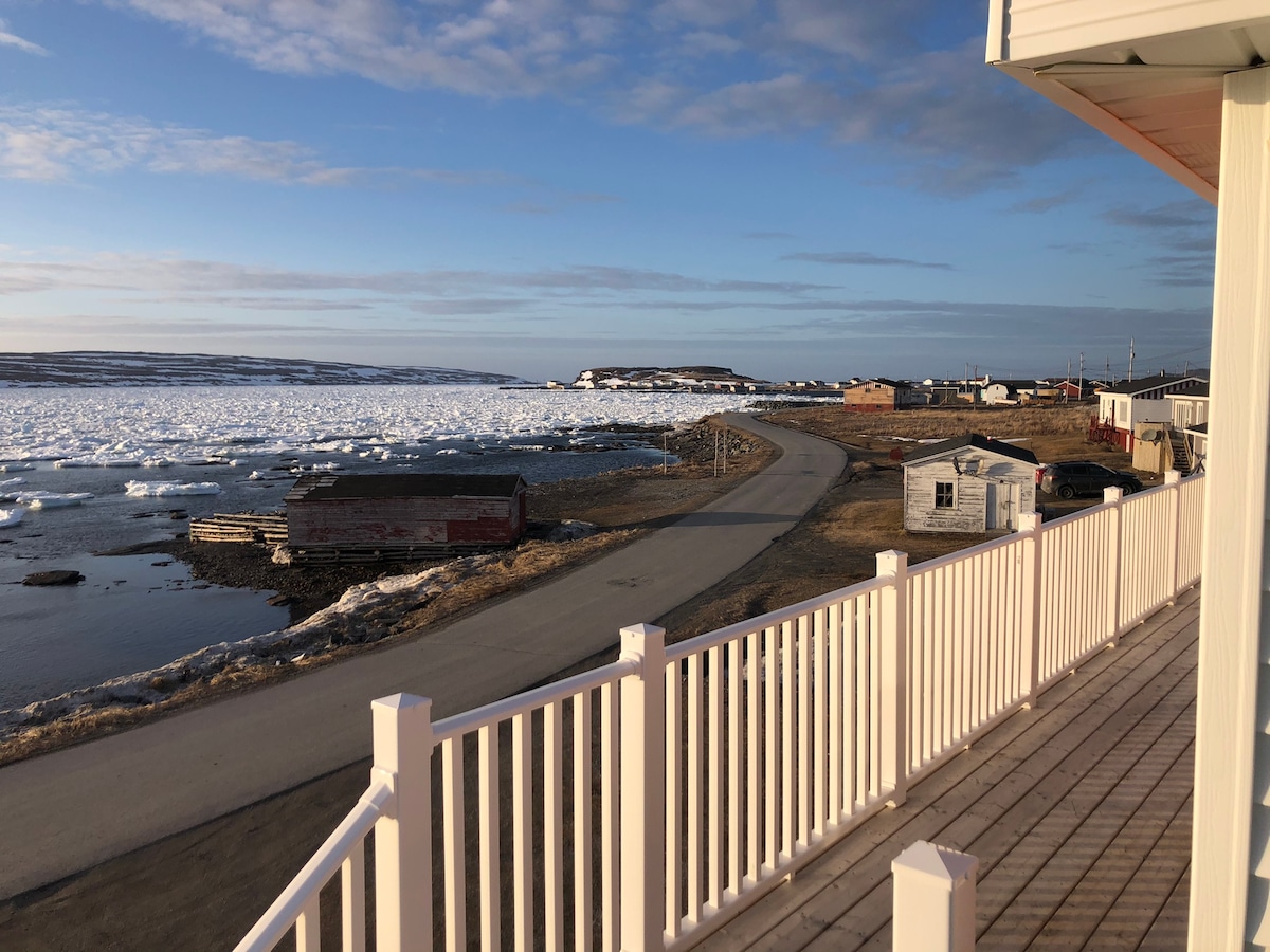 A scenic view is captured from a balcony, showcasing a winding road that runs alongside a tranquil waterfront. Ice and snow cover parts of the bay, while quaint buildings dot the landscape against a backdrop of a clear blue sky.