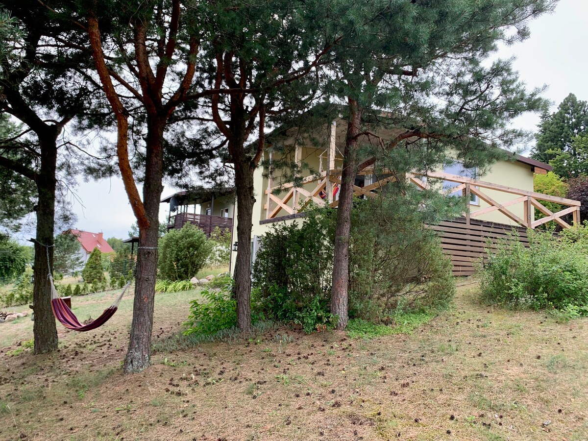 A glimpse of the house is framed by tall trees, providing shade and a natural setting. A hammock hangs between the trees, suggesting a peaceful spot for relaxation. The property features a grassy area surrounded by greenery, with the house visible in the background.
