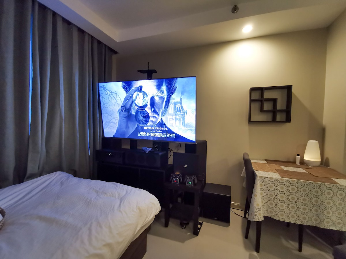 A spacious area is showcased with a large 65-inch TV prominently displaying a movie scene. Below, a sound system is positioned alongside a sleek console. A cozy bed sits nearby, and a dining table with a patterned cloth is visible, enhancing the living space.