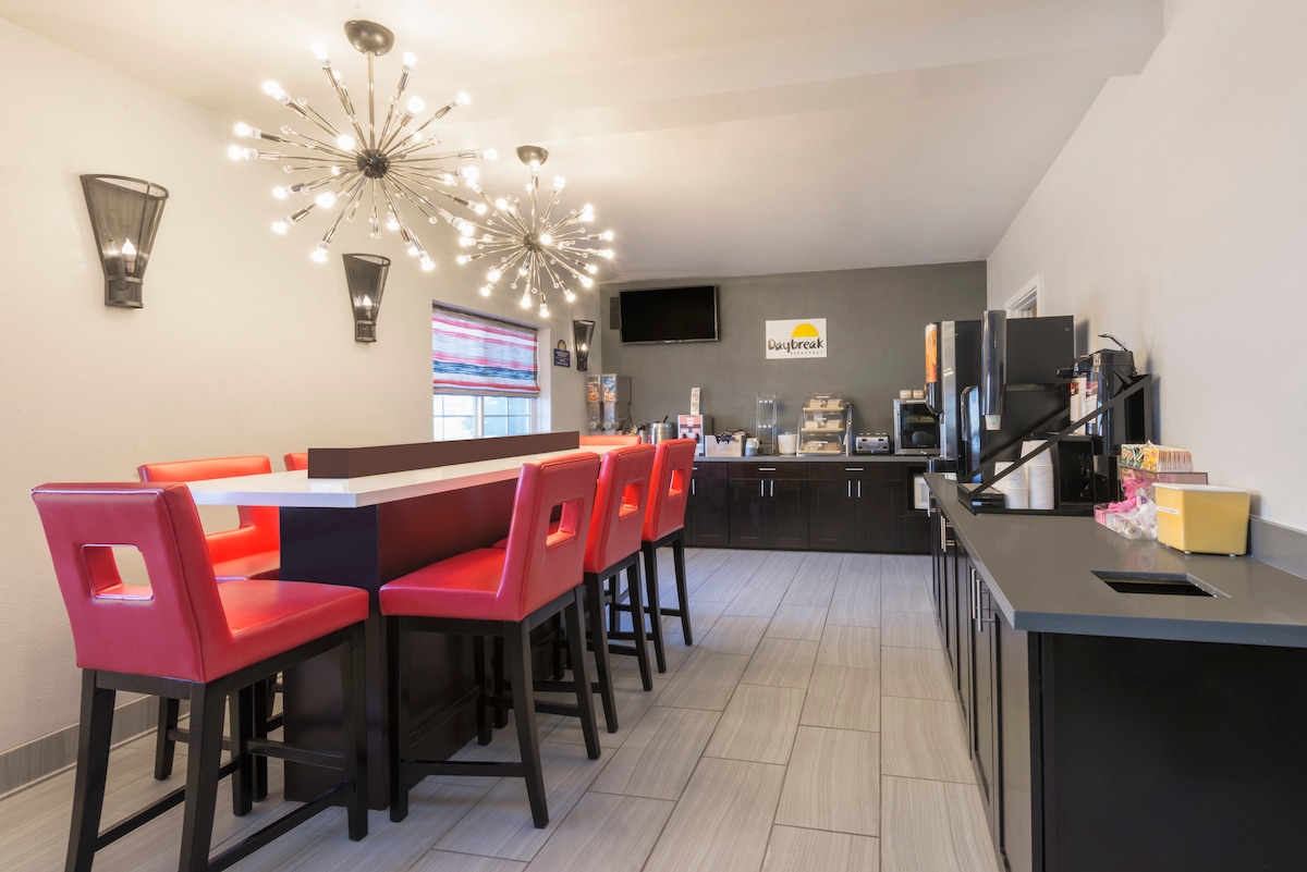 A modern dining area features a long table surrounded by high-backed chairs in bright red. A statement light fixture with multiple bulbs illuminates the space, while gray tile flooring adds a contemporary touch. A beverage station and neatly organized food options are visible in the background.
