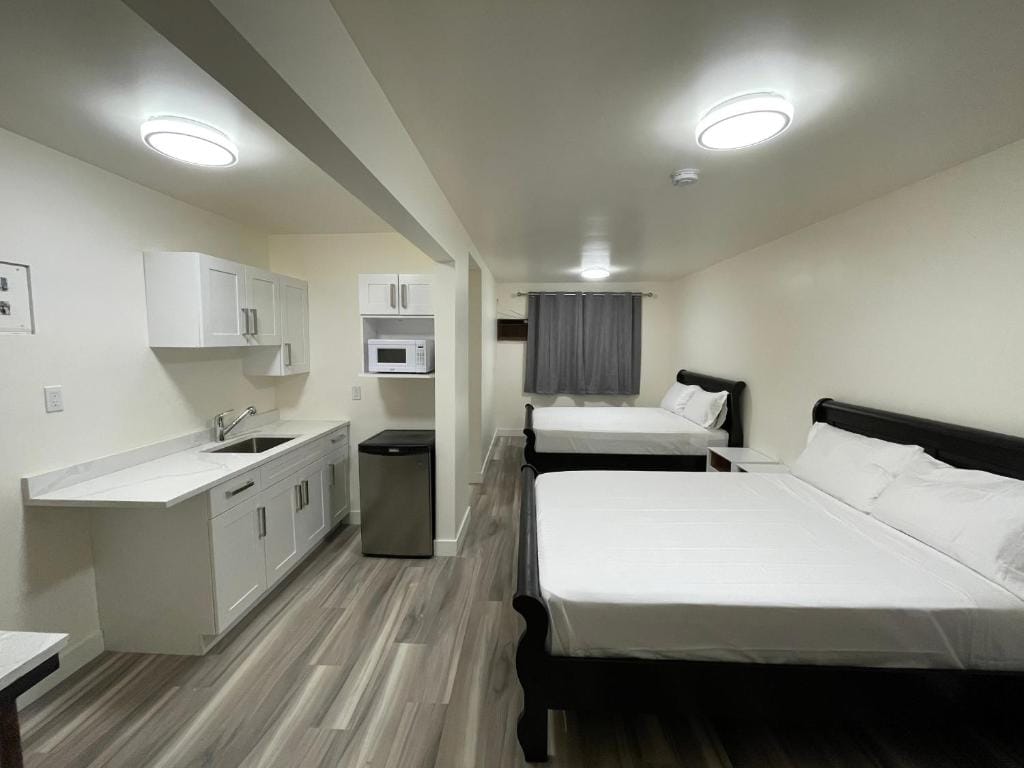 The suite features two queen size beds positioned opposite each other, with light-colored bedding. A kitchenette with modern appliances, including a microwave and mini fridge, is located along one wall. Neutral walls and flooring enhance the overall brightness of the space.