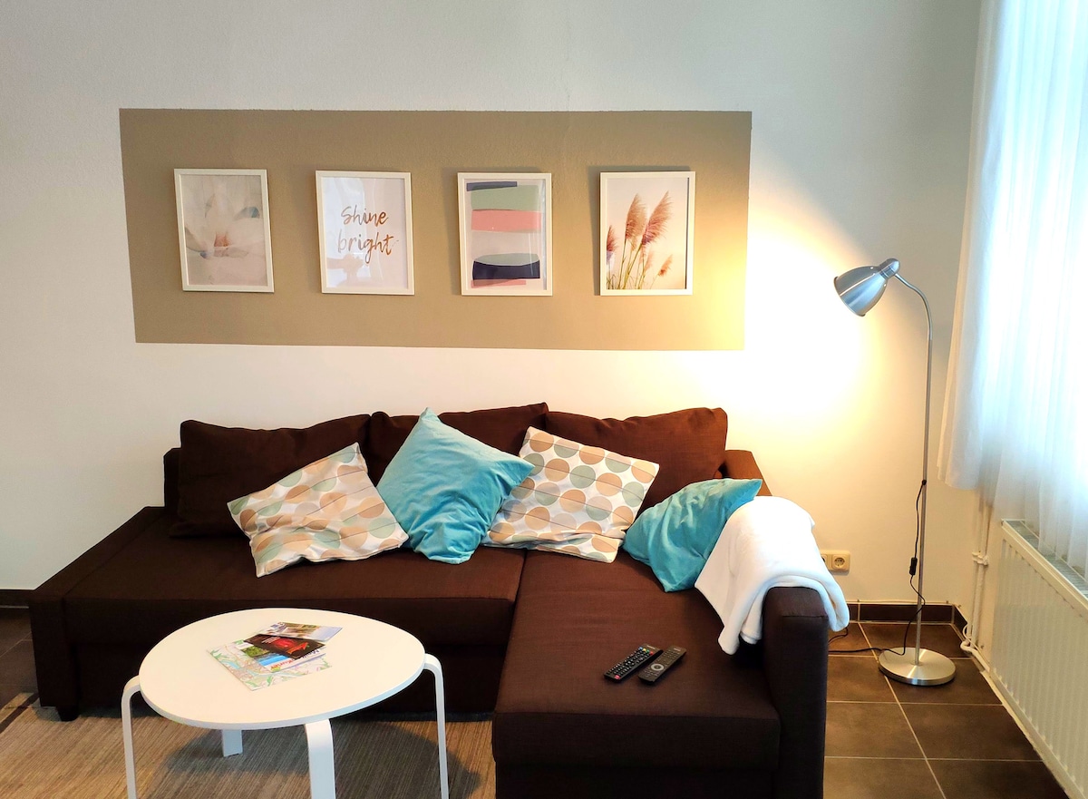 A modern living area is presented with a dark brown sectional sofa, adorned with colorful cushions featuring various patterns. Above the sofa, framed artwork adds a decorative touch, while a sleek floor lamp provides additional lighting. A round white coffee table sits in front of the sofa.