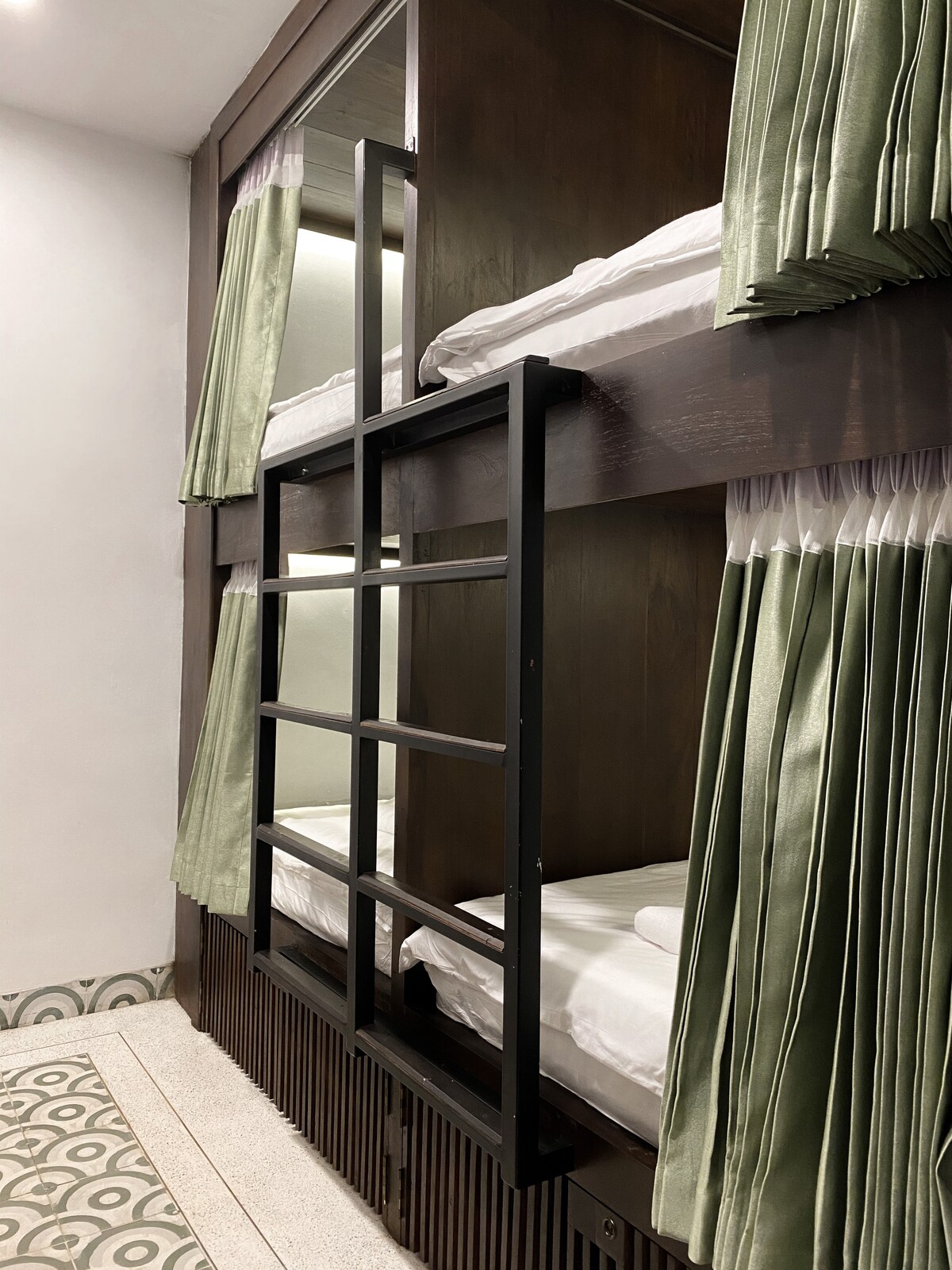 A pair of modern bunk beds is featured, showcasing an elegant wooden finish. Each bed is adorned with soft white linens and green curtains that provide privacy. A sturdy black ladder leads to the top bunk, while patterned flooring adds a decorative touch to the space.