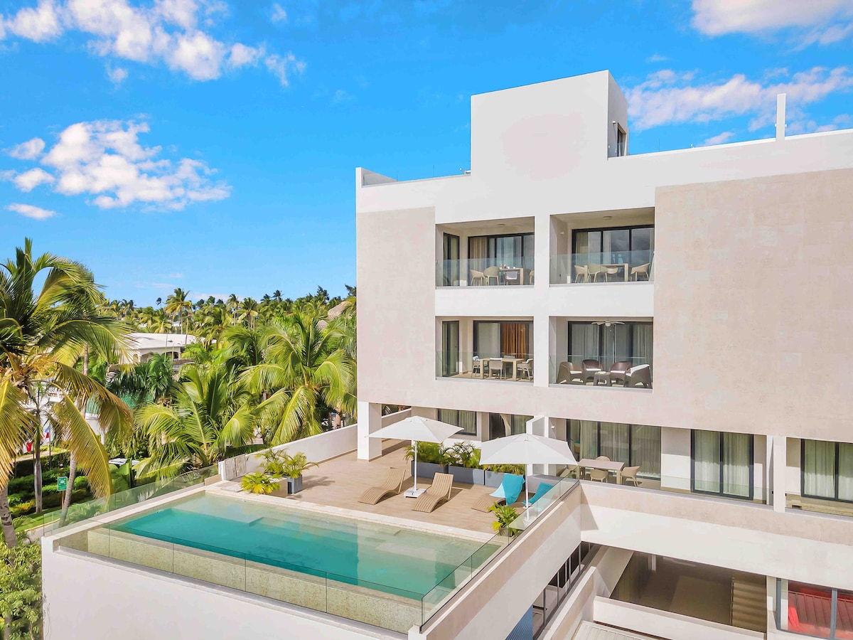 A modern apartment building is showcased, featuring a private terrace with an infinity pool. Sun loungers and umbrellas are positioned for comfort. Lush palm trees surround the area, with clear blue skies visible above the structure.