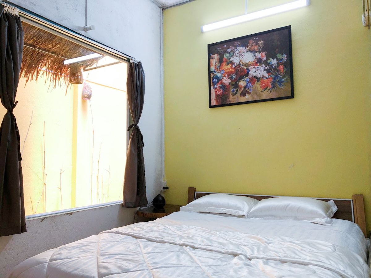A cozy sleeping area features a spacious bed with white linens, complemented by a colorful wall art piece above. Natural light enters through a window adorned with dark curtains, illuminating the warm yellow walls and providing a relaxed ambiance.