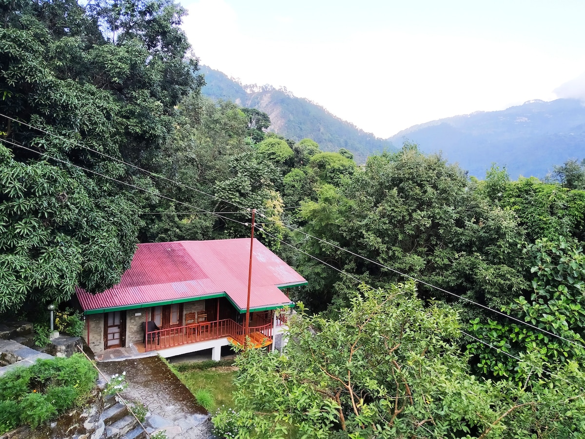 A cottage with a red roof is nestled among lush greenery, surrounded by trees. The foothills of the Himalayas provide a scenic backdrop, with soft clouds hovering above. A stone pathway leads to the entrance, enhancing the natural setting.