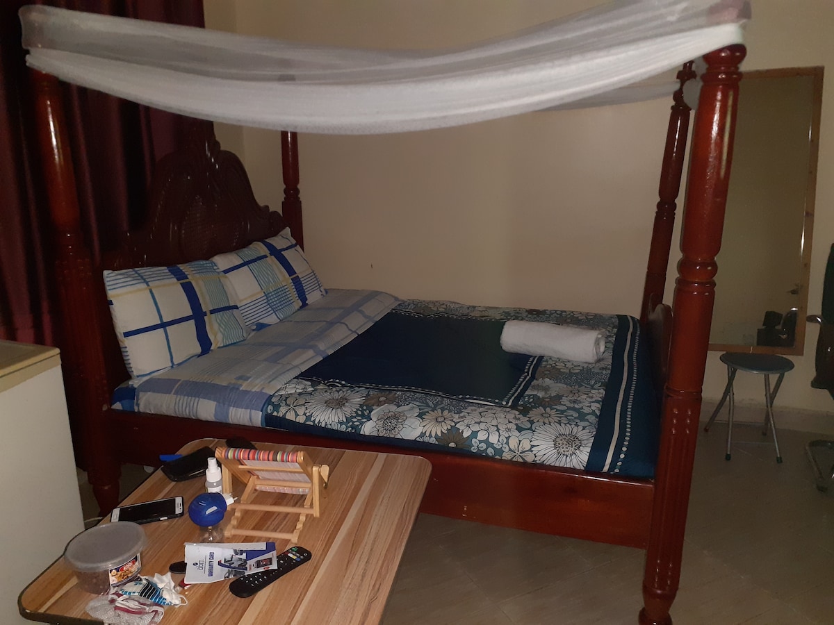 A spacious bed with a decorative patterned duvet is framed by a wooden canopy. A towel is neatly placed at the foot of the bed. A small table with various items and a chair are positioned nearby, complemented by soft lighting from the window draped with sheer curtains.