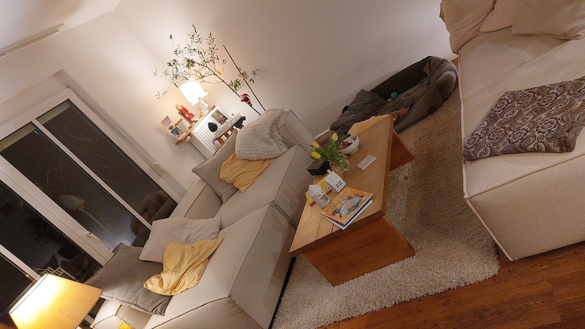 A comfortable living area features a light-colored sectional sofa adorned with throw pillows and blankets. A wooden coffee table sits in the center, surrounded by decorative items and a vase with flowers. Soft lighting enhances the space, with large windows allowing natural light.