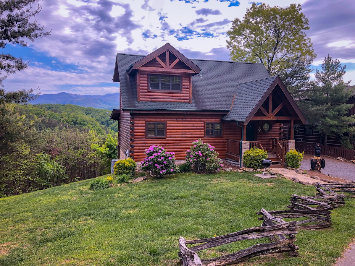 A wooden cabin is viewed from the front, surrounded by lush green grass and colorful flowering shrubs. The structure features a peaked roof and large windows that offer glimpses of the scenic mountains in the background under a partly cloudy sky.