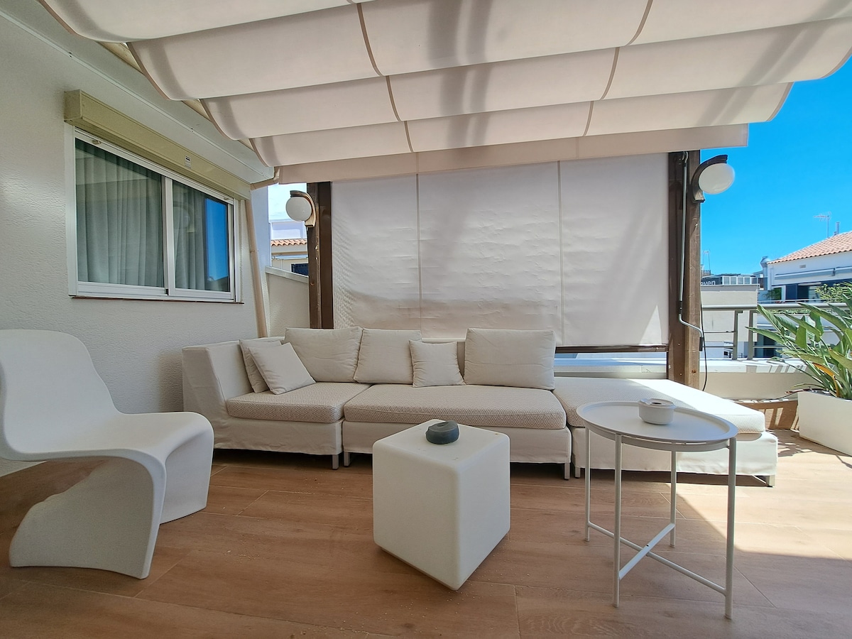 A bright terrace area is shown, featuring a comfortable outdoor seating arrangement with soft cushions. A small white table is positioned nearby, enhancing the inviting ambiance. Large windows allow natural light to fill the space, and a retractable awning provides shade.